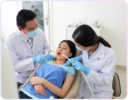 Dua dokter gigi dewasa (pria dan wanita) sedang melakukan perawatan gigi pada pasien anak perempuan di kursi dental klinik gigi.