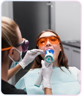 Veneer Gigi 3 Dokter gigi sedang melakukan prosedur pemutihan atau pemasangan veneer gigi pada pasien wanita yang memakai kacamata pelindung oranye dengan lampu curing, menunjukkan perawatan estetika gigi.
